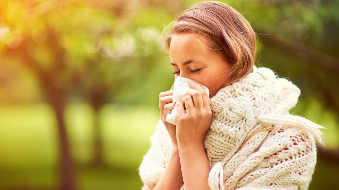 Femme avec pull d'hiver blanc qui se mouche à cause d'allergie, arrière plan flou d'arbres
