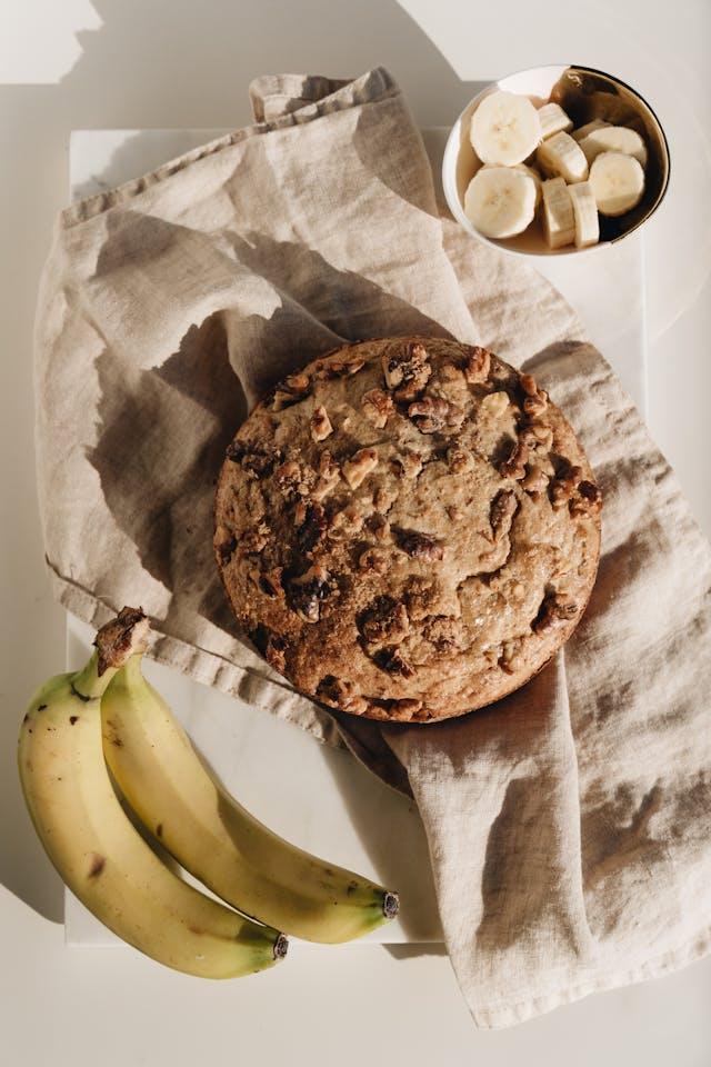 Deux bananes entières avec peau avec au milieu gâteau au noix posé sur un torchon beige et au fond des bananes en rondelles dans ramequin blanc