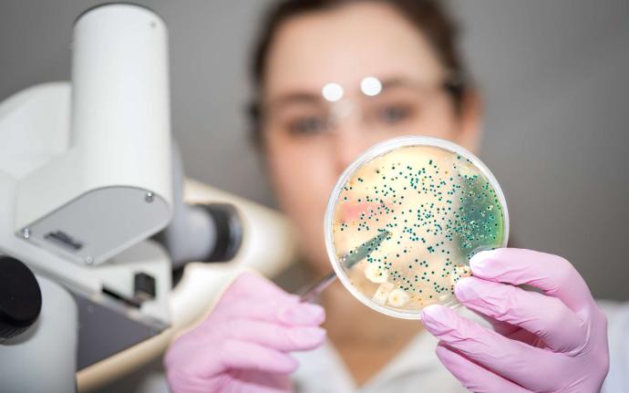 Visage femme floue en blouse blanche qui tient un rond transparent ave des petites billes vertes dedans et microscope blanc au premier plan à gauche
