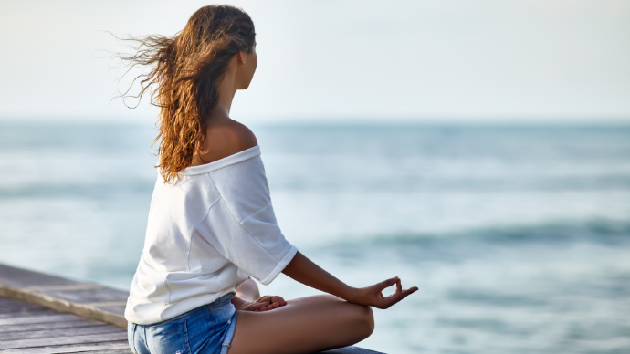 Jeune femme trois quart avec tee-shirt blanc et short en jean en tailleur position yoga face à la mer