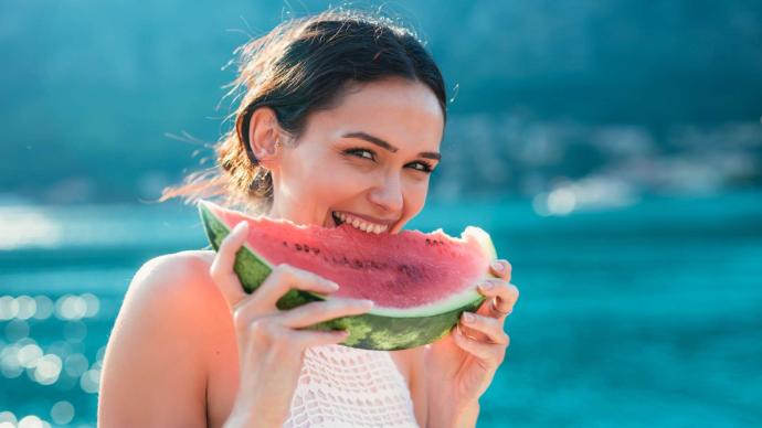 Jeune femme brune avec débardeur blanc qui mange un pastèque en souriant, arrière-plan flou mer turquoise