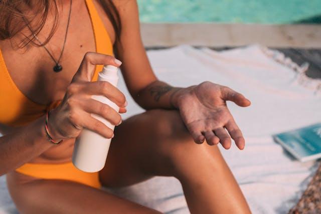 Femme avec maillot de bain une pièce orange assise sur serviette blanche au bord d'une piscine qui tient un flacon blanc de crème solaire