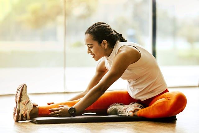 Jeune femme avec une tresse haut balnc legging orange et baskets qui s'étire devant une baie vitrée