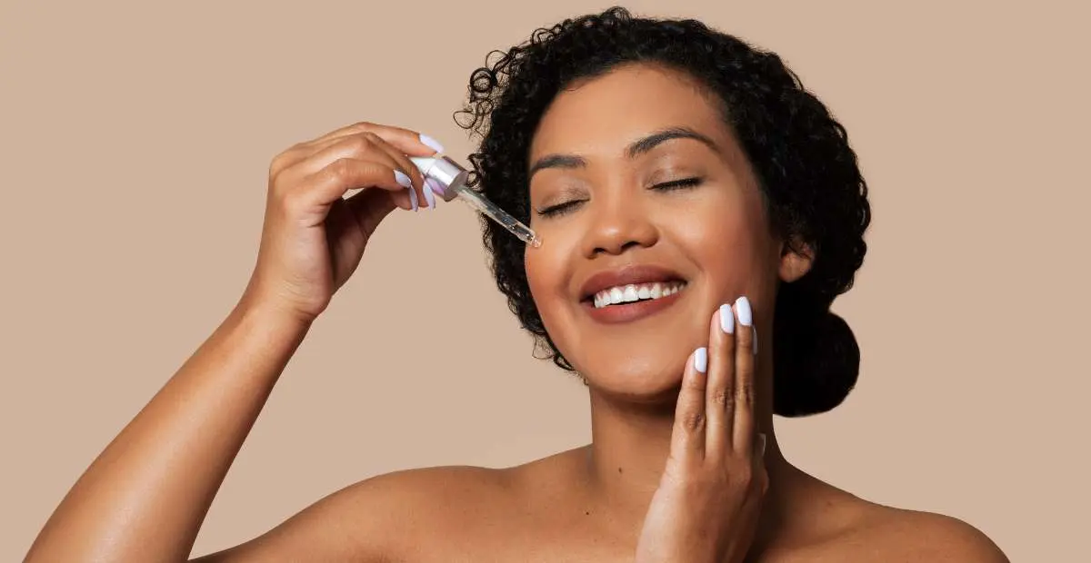 Jeune femme noire qui sourit les yeux fermés et qui tient une pipette de sérum posée sur sa joue