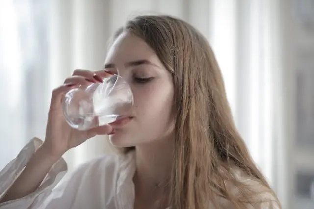 Jeune femme aux cheveux longs en chemise blanche de trois-quart qui boit un verre d'eau avec voilages blancs en arrière-plan